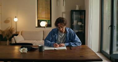A man with glasses is writing in a notebook sitting at a table with a serious face. Crumpled paper on the table. Stylish home interior photo