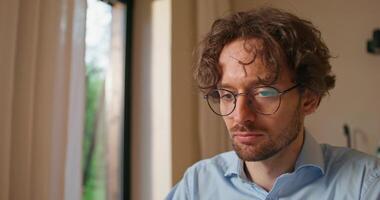 A man with glasses uses a laptop sitting at a table at home. Close-up of his focused face photo