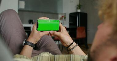 A man holds a mobile phone with a green screen lying on the couch at home. Close-up of his hands, an unrecognizable person photo