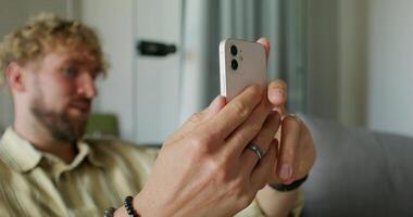 A bearded man in casual clothes is talking on a chat on a mobile phone sitting on the couch at home. The focuses on his hands. Daylight photo