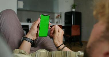 A man uses a mobile phone with a green screen lying on the couch at home. Close-up of his hands, an unrecognizable person photo