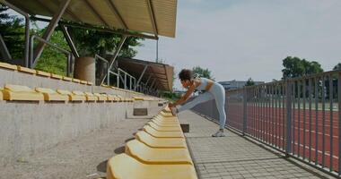 A woman in sports clothes and with earbuds in her ears does exercises standing near the seats at the city stadium on a summer day photo