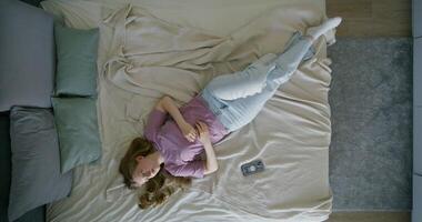 A young woman in casual clothes is lying on a bed during the day. The mobile phone is next to her. Top view, daylight photo