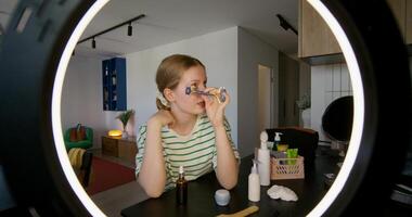 A young woman is doing a facial massage using a roller massager. She's talking while looking at the camera. through a round lamp photo