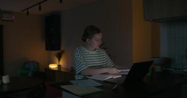A woman is reading a book and writing in a notebook while sitting at a table in front of an open laptop. Dark room, home interior photo