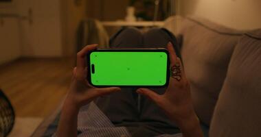 A woman uses a mobile phone with a green screen lying on the couch at home, close-up of her hands, an unrecognizable person, dimmed evening light photo