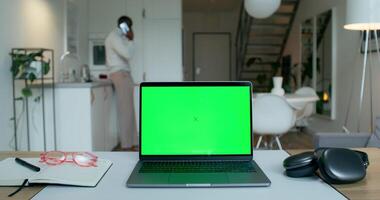 A laptop with a green screen is on the desktop at home. A man is talking on a mobile phone standing in the kitchen in the background photo