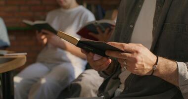 A man and a woman hold Bibles in their hands, sitting at a table indoors, close-up of their hands, unrecognizable persons. Daylight photo