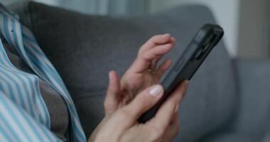 A woman in casual clothes uses a mobile phone while sitting on the couch at home. Close-up of her hands, an unrecognizable person photo