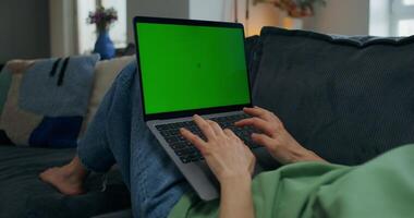 A woman typing on a laptop with green screen lying on the couch at home, close-up of her hands, an unrecognizable person photo