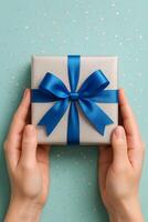 Gift Wrapped in Silver With a Blue Ribbon Held in Hands Against a Light Blue Pastel Background photo