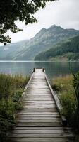 Tranquil Wooden Dock Extends Over Calm Lake Surrounded by Mountains on a Cloudy Day. photo