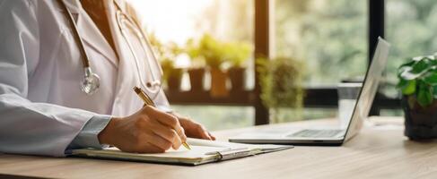 The doctor writing notes at a desk with a laptop and greenery in the background. photo