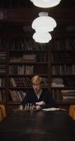 A young man of European appearance, dressed in a business suit, is studying with concentration to play chess alone, using a textbook, sitting at a table in the library, vertical shooting - business photo