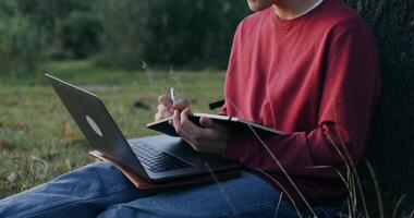 A young man of European appearance in a knitted hat, uses a laptop and writes in a notebook while sitting on the ground under a tree. - knitted photo