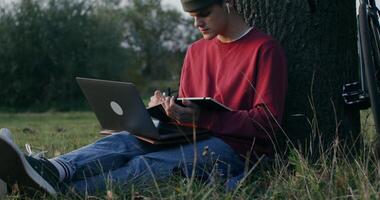 A young man of European appearance in a knitted hat, uses a laptop and writes in a notebook while sitting on the ground under a tree. photo