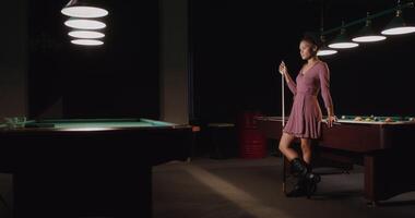 A beautiful young African-American woman, dressed in a dress, with a cue in her hands, stands leaning on a billiard table and smiles while looking at the camera - stands photo