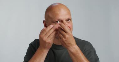 A bald man puts patches under his eyes. He smiles at the camera while standing in the studio background photo