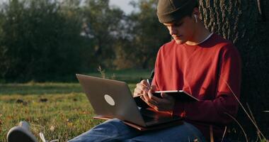 A young man of European appearance in a knitted hat, uses a laptop and writes in a notebook while sitting on the ground under a tree. - laptop photo