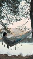 Peaceful freelancer working remotely on laptop while relaxing in hammock, surrounded by serene winter forest and frozen lake landscape, embodying digital nomad lifestyle and work-life balance video