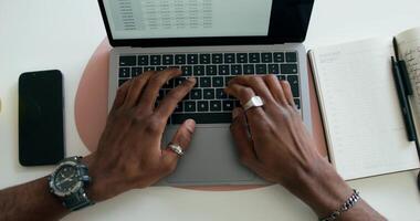 A man typing on a laptop, sitting at a table indoors, close-up of his hands, an unrecognizable person, view from above. An open notebook is on the table - notebook photo
