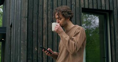 A man in casual clothes uses a mobile phone and drinks coffee standing on the terrace on a summer day. photo