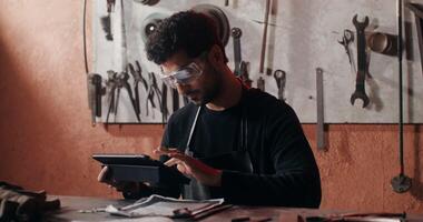 A young blacksmith in goggles and an apron uses a tablet while sketching a future product, sitting at a desk in a forge full of working tools. in 4k, red komodo photo