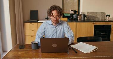 A man with glasses works from home. He uses a laptop and drinks coffee sitting at the kitchen table photo