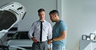 A car dealership manager talks to a customer standing next to a car with the hood open in the showroom photo