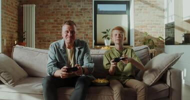Father and son play games with joysticks and eat popcorn sitting on the couch at home, happy childhood - Father photo