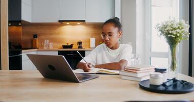 The girl does her homework at home, she uses a laptop and makes notes in a notebook while sitting at the kitchen table - notebook photo