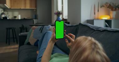 A woman uses a mobile phone with green screen while lying on the couch at home. A rear view of her hands, an unrecognizable person photo