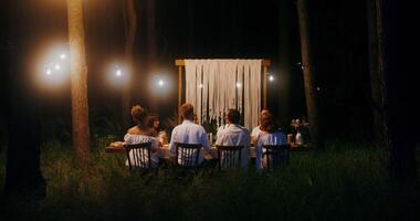 The bride and groom kiss standing up from behind the covered festive table, at which the guests are sitting - festive photo