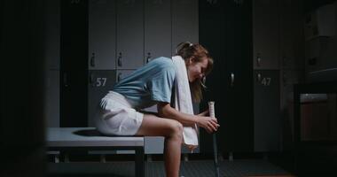 Young beautiful woman tennis player is resting in the locker room after the match, sitting on a bench with a towel around her neck and twirling a tennis racket in her hands. photo