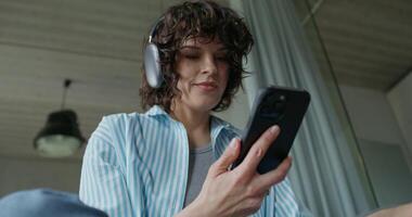 A woman in casual clothes and with headphones is using a mobile phone and smiling. She's sitting on the couch at home. Daylight - headphones photo