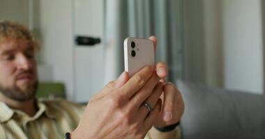 A bearded man in casual clothes is talking on a chat on a mobile phone sitting on the couch at home. Daylight photo