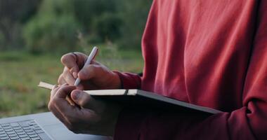A young man of European appearance makes notes in a notebook, using a laptop, sitting on the ground under a tree, close-up photo