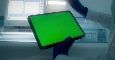 A woman in a lab coat and disposable gloves holds a tablet with a green screen standing in a medical laboratory. Close-up of her hands, copy space - screen photo