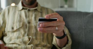 A man in casual clothes uses a tablet sitting on the couch at home. He enters credit card details for online purchases. Daylight photo