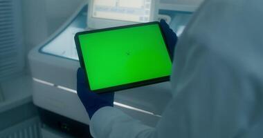 A woman in a lab coat and disposable gloves holds a tablet with a green screen standing in a medical laboratory. Close-up of her hands, copy space - tablet photo