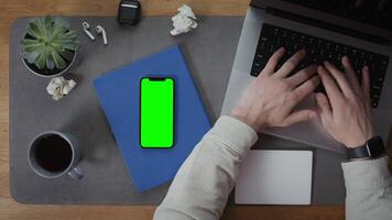 A man is typing on a laptop keyboard, sitting at a desktop littered with papers, headphones and a mobile phone with a green screen lying next to it, close-up. Mockup, chroma key, copy space - sitting photo