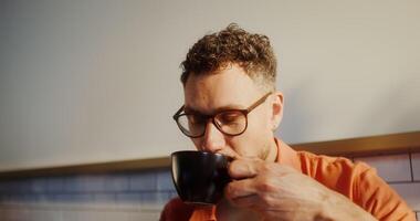 A stylish young man with glasses drinks coffee sitting in a cafe, face close photo