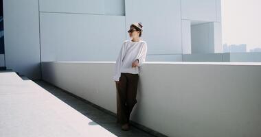 A young woman of European appearance in sunglasses stands on the balcony of a white minimalist building and looks around - stands photo