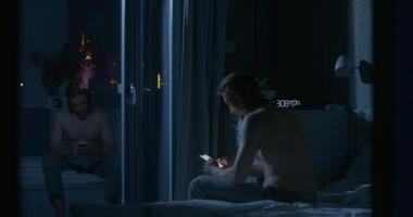 A man in pajama pants uses a mobile phone while sitting on the bed at night. Dark room, home interior - sitting - in photo