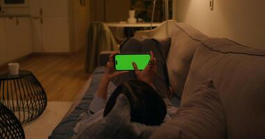 A woman uses a mobile phone with a green screen lying on the couch at home, dimmed evening light - dimmed photo