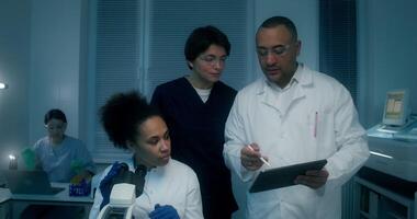 A group of laboratory technicians perform a blood test in the laboratory. A man and a woman use a tablet standing near a laboratory assistant's desk with a microscope - standing photo