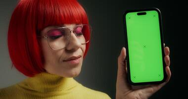 A woman with glasses and an orange wig smiles at the camera, holding a mobile phone with green screen in her hand, standing on a gray background in the studio - holding photo