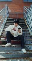 A young man of Chinese appearance uses a mobile phone and, drinks juice from a disposable cup with a straw while sitting on an outdoor metal staircase photo