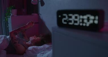 A woman uses a mobile phone lying on the bed. Dark room, home interior. The electronic clock is on the bedside table - bedside photo