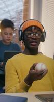 A man and a woman use laptops working at desks in the office. A man in a hat and headphones is chatting on chat and playing with a baseball photo
